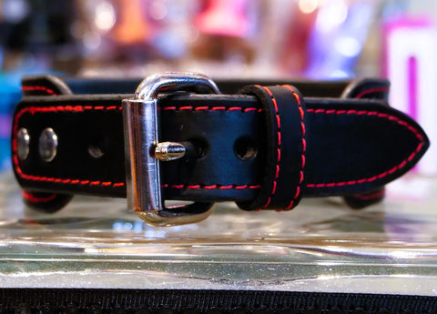 A close-up of a black leather collar with vibrant red contrast stitching and a polished silver-tone buckle, resting on a reflective glass surface.