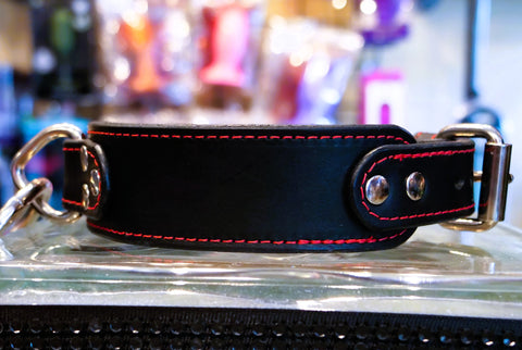 A black leather collar with red contrast stitching and silver-tone metal hardware, including a buckle, D-ring, and adjustment rivets, resting on a clear glass surface.
