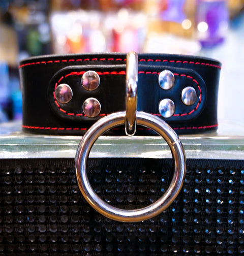 A black leather collar with red stitching, silver studs, and a large polished silver metal O-ring, displayed on a reflective surface.