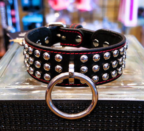 A black leather collar adorned with silver-tone studs and red stitching, featuring a large polished metal O-ring, displayed on a clear acrylic stand.