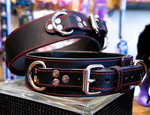 A black leather thigh cuff with red contrast stitching and silver-tone hardware, displayed on a clear acrylic stand in a store setting.