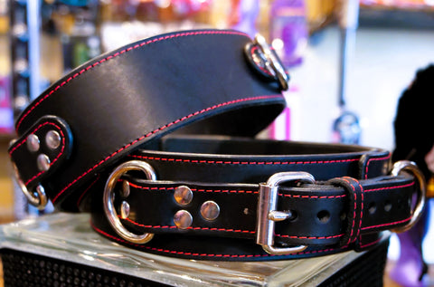A close-up of a black leather thigh cuffs with red contrast stitching and silver-tone metal hardware, including a buckle, rings, and studs, displayed on a clear stand.