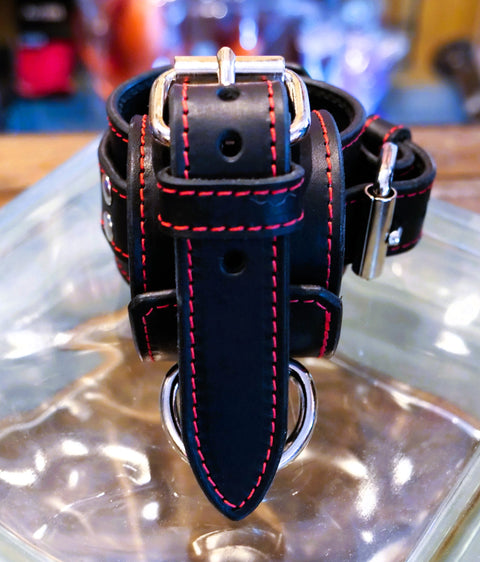 A black wrist cuff with vibrant red contrast stitching and silver-tone metal hardware, displayed on a clear glass surface.