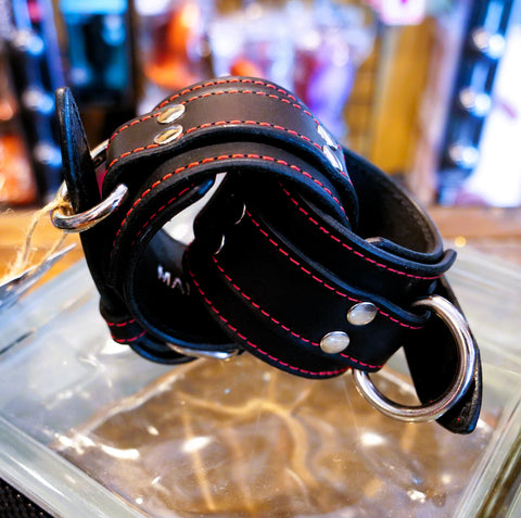 A black leather wrist cuff with red contrast stitching and silver-tone metal hardware, including a large D-ring and rivets, resting on a clear glass surface with a blurred background.