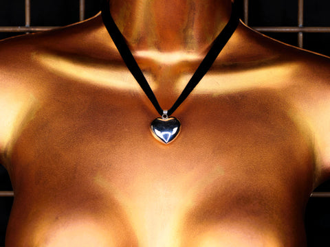 Heart-shaped silver bell pendant on a black velvet cord against a bronze-colored surface.
