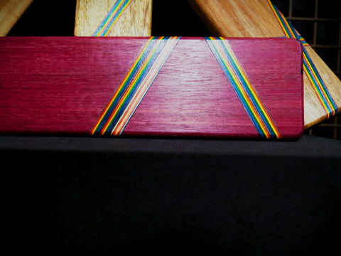 A close-up of a rectangular wooden board with a deep reddish-purple finish and two diagonal bands of multicolored wood grain, resting on a black surface.