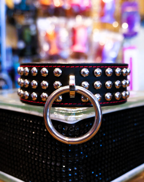 A black studded collar with a large silver metal O-ring, displayed on a black textured surface with a blurred shop background.